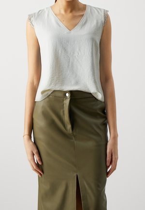 Femme portant un haut blanc sans manches bordé de dentelle, rentré dans une jupe en cuir vert olive taille haute avec une fente à l'avant et une fermeture à boutons.