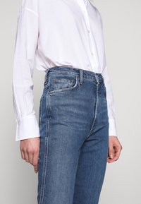 Jeans de mezclilla azul de tiro alto con corte recto, con herrajes en tono cobre y discretos desgastes, combinados con una camisa blanca de botones.