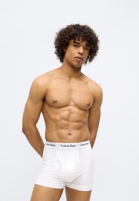 White cotton boxer briefs with a contrasting black Calvin Klein waistband, fitted design, and soft texture. Model displays a neutral pose.