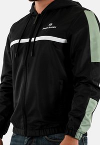 Giacca nera con cappuccio, dettagli in bianco e verde chiaro, caratterizzata da chiusura con zip, polsini e bordo elasticizzati, e un tessuto testurizzato.