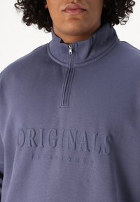 Suéter azul con cuello alto y cremallera, con el texto en relieve "ORIGINALS JACK & JONES." Tejido suave con una textura lisa.