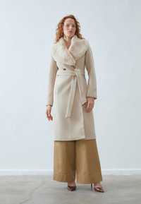 Beige wool coat with a large faux fur collar, double-breasted buttons, and a belted waist, paired with wide-leg khaki pants and heels.