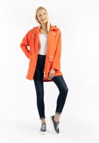 Egy nő mosolyog, narancssárga esőkabátban, fehér ujjatlan felsőben, sötét, szűk farmert viselve, kék sportcipőben áll egy fehér háttér előtt.