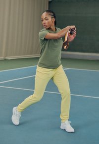 En person spiller tennis, iført en grønn skjorte og gule joggebukser, med et tennisracket i hånden, mot en blå tennisbane.