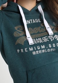 Felpa color turchese con cordini bianchi, caratterizzata da testo stampato in oro e argento. Tessuto morbido con tasca frontale e design classico.