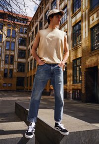 Beige mouwloze t-shirt, blauwe jeans met een versleten patch, zwart-witte sneakers en een bijpassende pet, tegen een bakstenen gebouw.