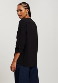 Czarna koszula z długim rękawem o luźnym kroju, z teksturowanej tkaniny i z zapinanymi rękawami. Połączona z luźnymi niebieskimi dżinsami.