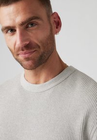 Gray ribbed sweater with a round neckline, textured fabric, and minimalist design. Model has an earring and light stubble.