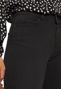 Vaqueros negros con un diseño clásico de cinco bolsillos, silueta ajustada y detalles de costura mínimos. La superficie es suave con un ligero estiramiento.
