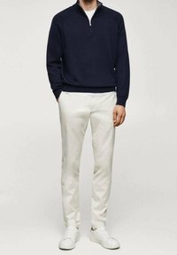 Un sweat à fermeture éclair bleu marine, un t-shirt blanc en dessous, un pantalon crème, et des baskets blanches. Design simple, texture lisse et détails de quincaillerie minimaux.