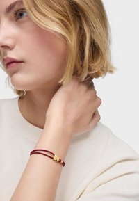 Pulsera de cuero rojo con un broche dorado que presenta un diseño de mariposa, llevada en una muñeca, sobre un fondo neutro claro.