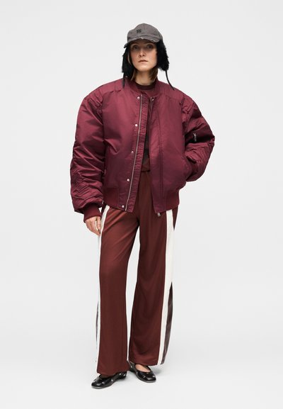 Veste bomber bordeaux avec poignets côtelés, pantalon large assorti avec des bandes latérales blanches et brun foncé, et une casquette grise avec des cache-oreilles.