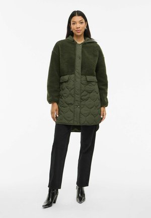 Manteau matelassé vert avec un dessus en velours texturé, deux poches avant, une capuche et des manches longues, associé à un pantalon noir à jambe droite et des bottes à talons.