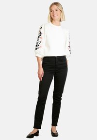 Donna in piedi che indossa una blusa bianca con ricami floreali neri e rosa sulle maniche, pantaloni neri attillati e scarpe nere con tacco basso.