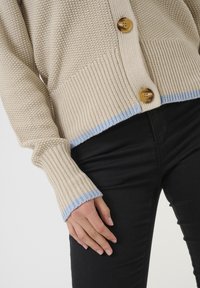 Beige knitted cardigan with textured pattern, featuring two large brown buttons and light blue accents at the hem and cuffs.