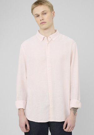 Jeune personne aux cheveux blonds courts portant une chemise rose clair à boutons avec manches retroussées, un pantalon foncé, et un tatouage visible sur le poignet gauche.