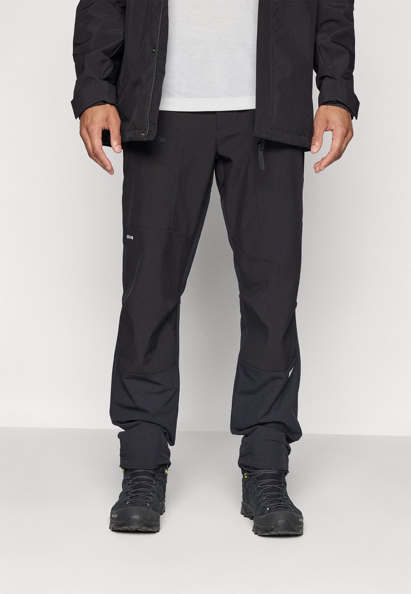 Homme portant un pantalon de plein air noir, des bottes de randonnée noires, une chemise blanche et une veste noire, debout devant un fond clair uni.
