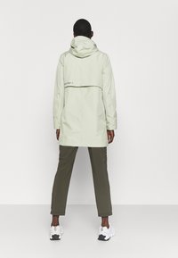 Ljusgrön vattentät jacka med huva, har en ryggklaff och subtil branding. Kombineras med olivgröna byxor och vita sneakers.