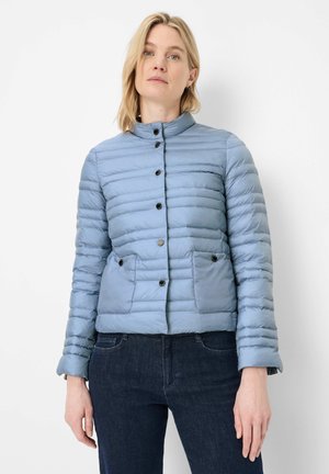 Frau trägt eine hellblaue gesteppte Jacke mit schwarzen Knöpfen, steht vor einem weißen Hintergrund, kombiniert mit dunkelblauen Jeans.