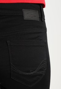 Jeans de denim negro con una etiqueta de cuero que muestra el nombre de la marca, un bolsillo trasero con costuras detalladas y un diseño curvado.