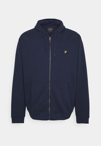 Sweat à zip bleu marine fabriqué en tissu doux, avec une poche kangourou et un petit emblème jaune sur le côté gauche de la poitrine.