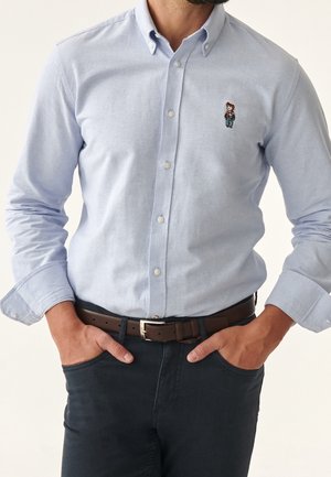 Hellblaues Hemd mit Knopfleiste und gesticktem Bärenlogo, dunkle Hose, brauner Gürtel, Hände in den Taschen, im Bildausschnitt am Hals und der Taille abgeschnitten.