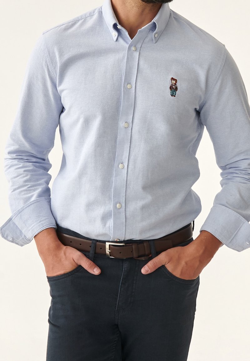 Chemise boutonnée bleu clair avec logo ourson brodé, pantalon foncé, ceinture marron, mains dans les poches, cadrée au niveau du cou et de la taille.