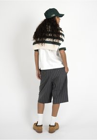 Femme portant une casquette verte, un pull rayé posé sur les épaules, une chemise blanche, un short gris à fines rayures, des chaussettes blanches et des chaussures marron, debout devant un fond blanc.