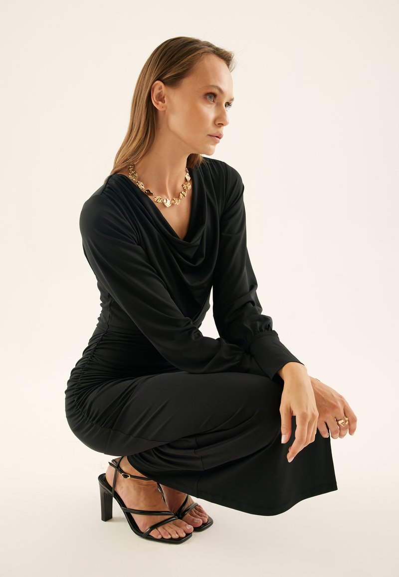Black, draped dress with long sleeves and ruched detailing. Paired with strappy black heels and a chunky gold necklace. Smooth fabric texture.