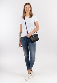 Bolso cruzado negro con superficie texturizada y detalles de cremallera, combinado con una camiseta blanca ajustada y jeans skinny azules descoloridos.