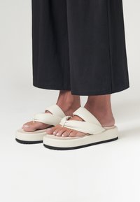 Infradito piattaforme bianche con un design a nodo sulle cinghie, caratterizzate da una suola testurizzata e una superficie liscia. Abbinati a pantaloni larghi scuri.
