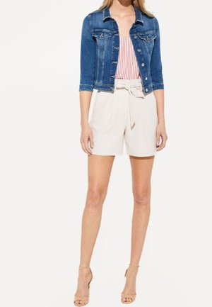 Kvinde iført en blå denimjakke, lyserød top med striber, højtaljede hvide shorts med et stofbælte og beige højhælede sandaler.