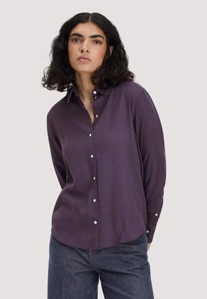 Frau mit lockigem dunklem Haar, die ein violettes Hemd mit Knöpfen und blaue Jeans vor einem schlichten hellen Hintergrund trägt.