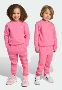 Dve deti v zladených jasne ružových mikinách a nohaviciach vyrobených z mäkkej látky, ozdobených bielymi logami Adidas. Tenisky dopĺňajú ich oblečenie.