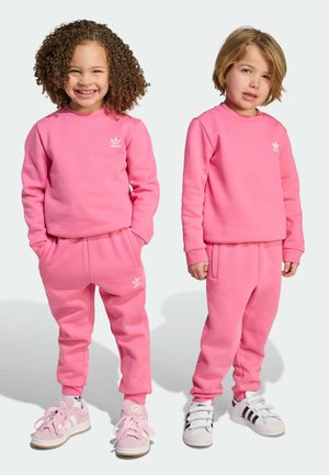 Két gyerek egymással megegyező, fényes rózsaszín szabadidőfelsőben és szabadidőnadrágban van, amely puha anyagból készült, és fehér Adidas logó díszíti. Edzőcipőik kiegészítik az öltözéküket.