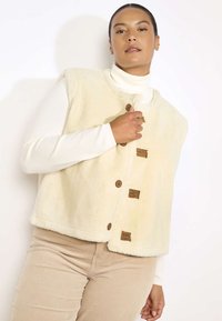 Krēma krāsas vilnas veste ar apaļu kakla izgriezumu, kurai ir trīs brūni pogu aizdares un gaiši brūni akcenti. Valkāta virs baltas apģērba ar augstu apkakli.