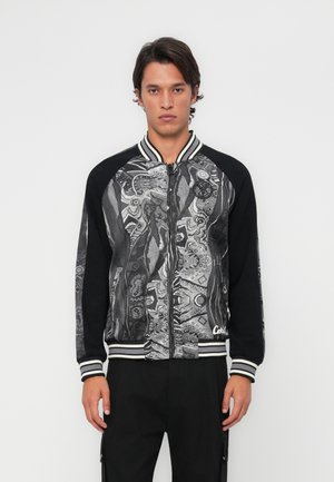 Black bomber jacket with patterned front, white accents, and ribbed cuffs. Features a zipper closure and graphic designs in monochrome.