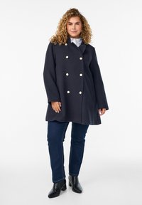 Navy double-breasted coat with large cream buttons, long sleeves, and a relaxed fit. Paired with dark blue jeans and black heeled boots.