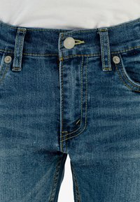Jeans de mezclilla en lavado azul oscuro con costuras amarillas. Las características incluyen cinco bolsillos, cierre de botón metálico y trabillas para el cinturón.