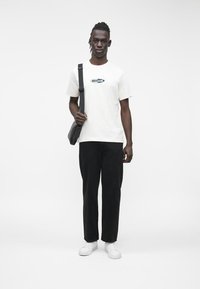 Mees, kes kannab valget "Guess Jeans" t-särki, musti pükse, valgeid tossusid ja musta ristihoidjat, seisab valge taustaga vastu.