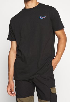 Man die een zwart T-shirt draagt met een klein blauw en roze logo op de borst en zwarte cargobroek met olijfgroene zakken.