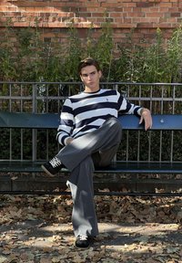 Jonge man in zwart-wit gestreepte trui en grijze broek zit met gekruiste benen op een parkbank, met gevallen bladeren en een bakstenen muur op de achtergrond.