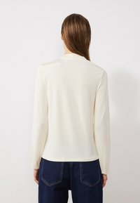 Langärmliges Oberteil in Off-White, aus glattem Stoff gefertigt. Verfügt über einen schlichten runden Halsausschnitt und ein tailliertes Design, kombiniert mit dunkelblauen Jeans.