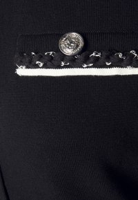 Tissu noir en tricot avec une rayure blanche contrastante, présentant une bande tressée décorative et un bouton en argent avec un design détaillé.