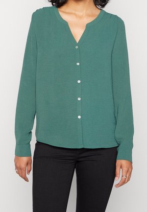 Femme portant un chemisier vert à manches longues boutonné et un pantalon noir, debout devant un fond clair uni.