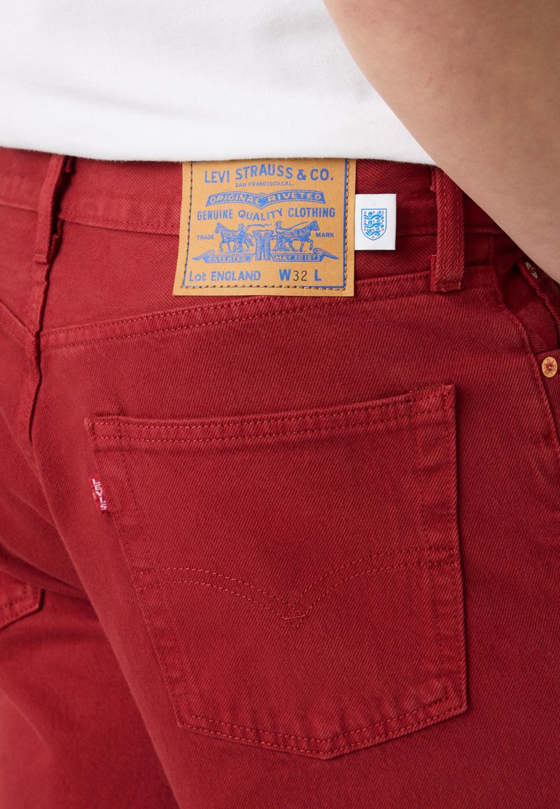 Bolsillo trasero rojo de unos vaqueros Levi's con una etiqueta de cuero Levi Strauss & Co. que muestra la talla de cintura W32 L, usados con una camisa blanca.