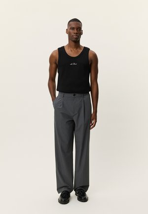 Débardeur noir sans manches avec logo, et pantalons larges gris à fermeture par bouton. Chaussures noires à lacets complètent la tenue.