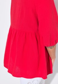 Top rosso tessuto con maniche a sbuffo corte, vita arricciata e orlo fluido. Presenta un tessuto leggero e stropicciato.
