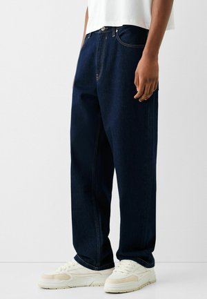 Person trägt dunkelblaue locker sitzende Jeans, weiße Sneakers und ein weißes kurzes Top, steht vor einem schlichten Hintergrund.