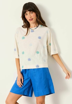 Kvinde iført en hvid kortærmet top med blå og grønne blomsterprint og lyseblå knælange shorts.